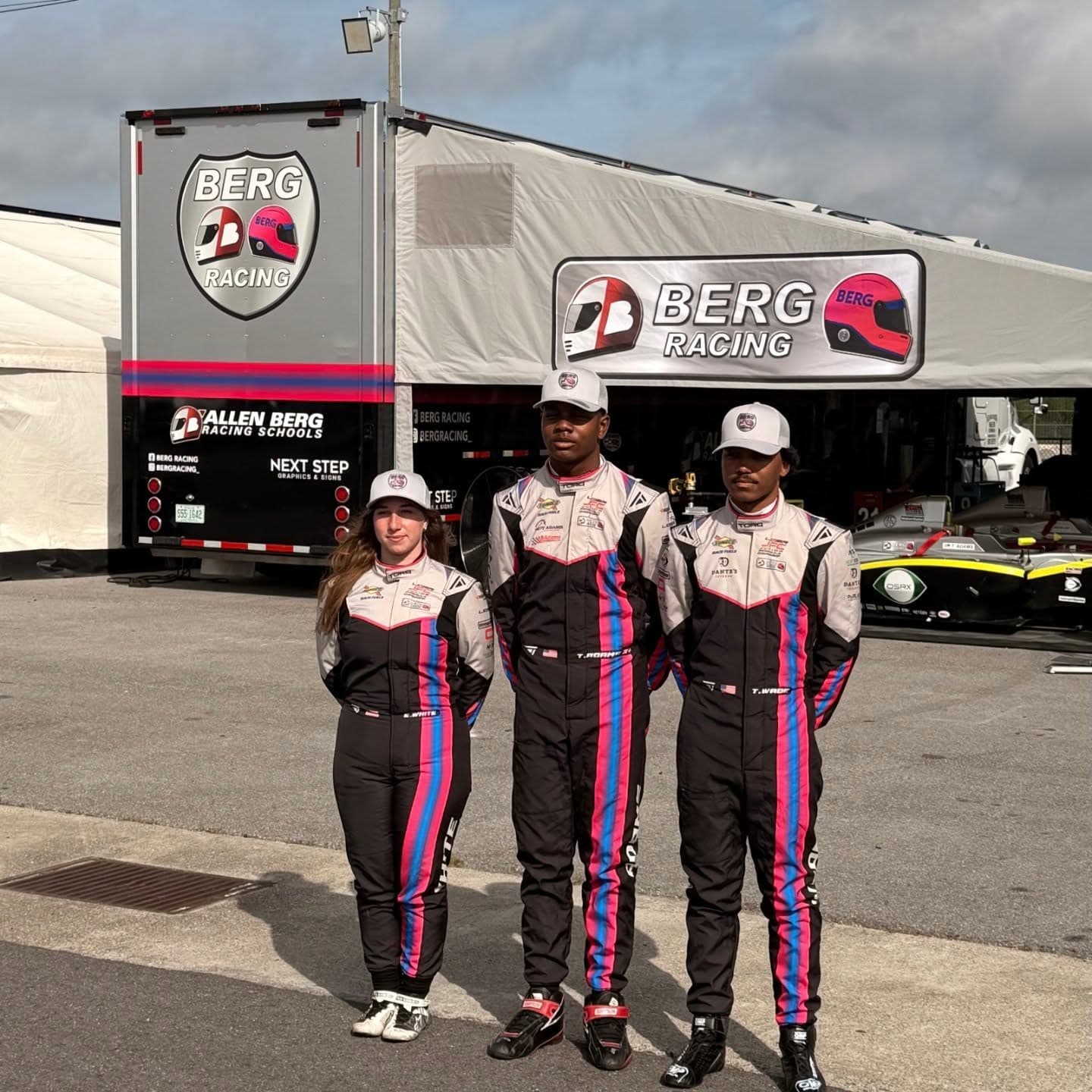 Three Berg Racing Drivers standing outside their awning in grey, pink and black Torq Racecraft suits.