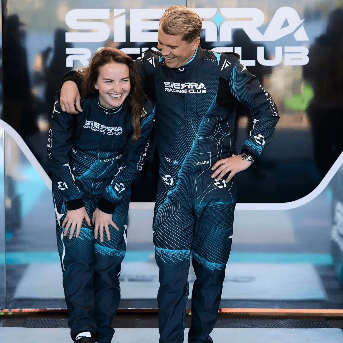 Two drivers laughing wearing black and teal Torq Racecrafts suits.