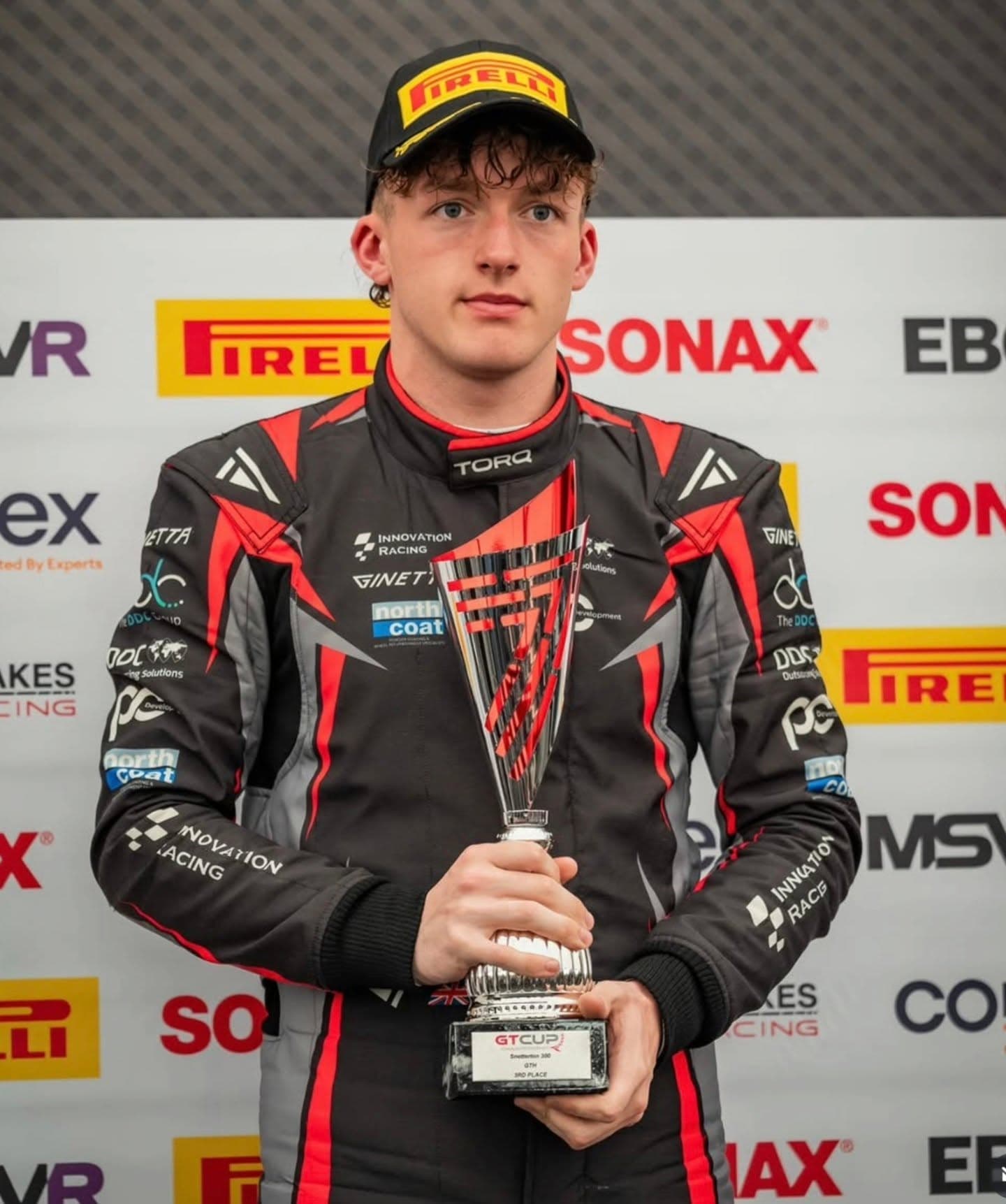James Wareing holding a trophy on the GT Cup podium wearing a black, grey and red Torq Racecraft