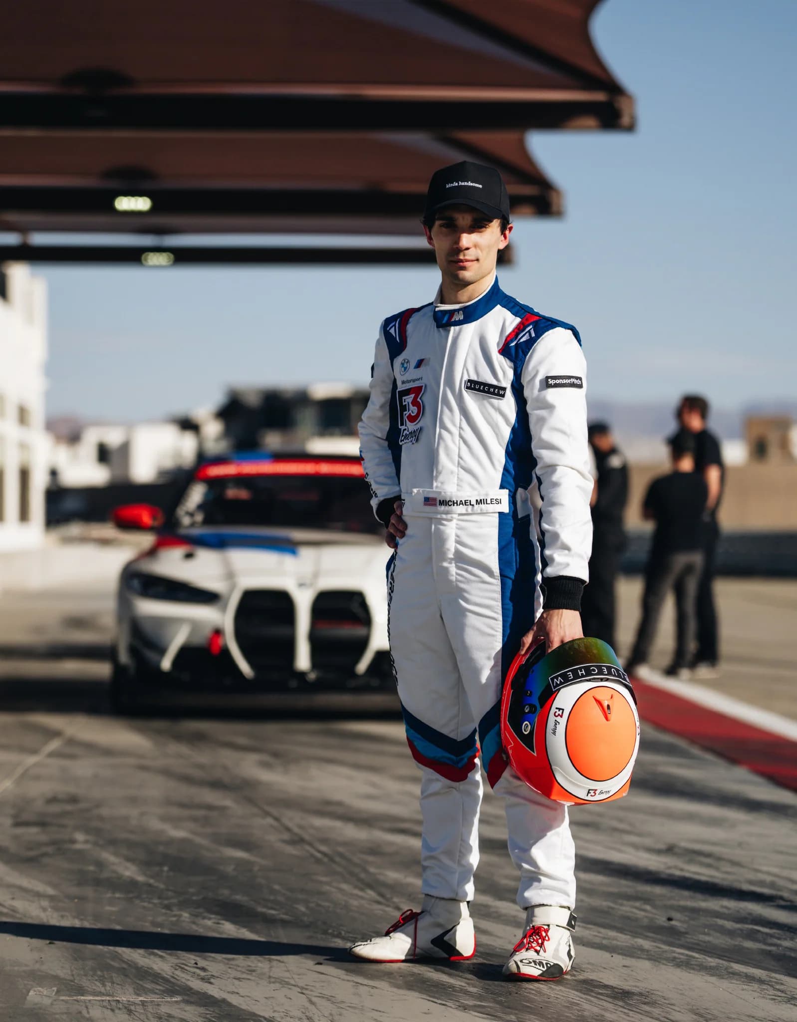 Driver in a white and blue Torq Superlight race suit holding a helmet in front of a BMW race car at the circuit.