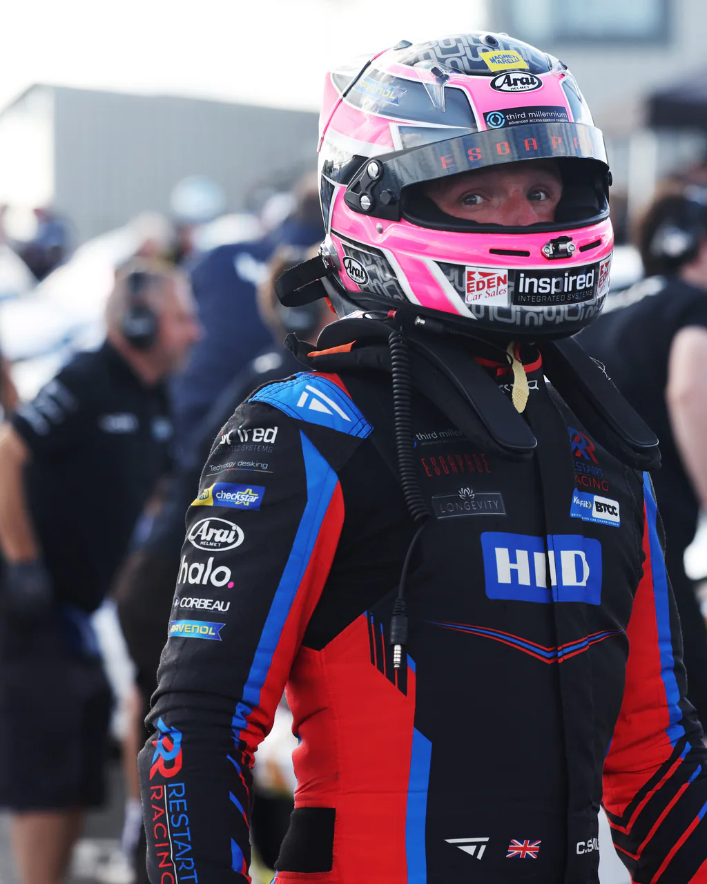 Helmeted racing driver in a black, red and blue Torq suit standing in a busy paddock.