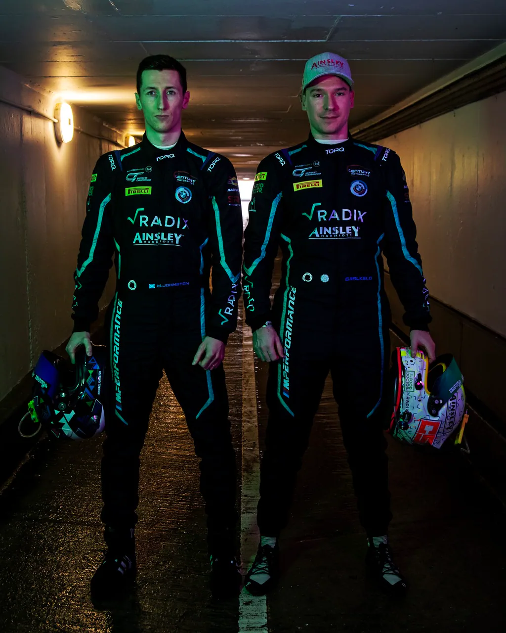 Two drivers wearing black and teal Torq race suits, holding their helmets in a tunnel.